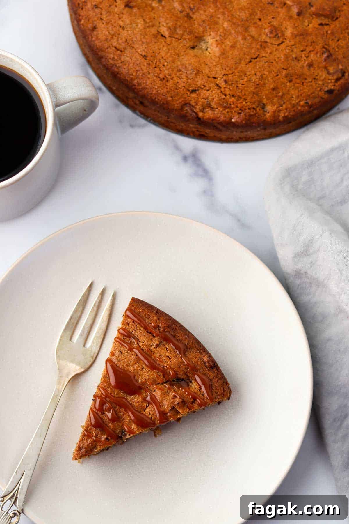 A slice of caramel-drizzled cake on a plate next to a fork and a cup of coffee.