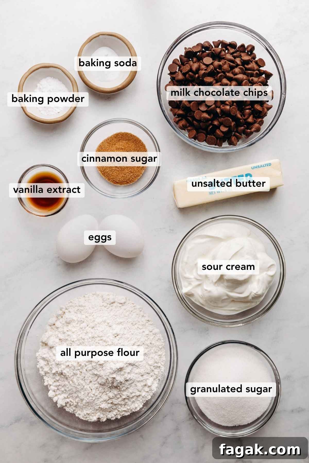 Ingredients in various size bowls labeled as baking soda, baking powder, milk chocolate chips, vanilla extract, cinnamon sugar, unsalted butter, eggs, sour cream, all purpose flour, and granulated sugar on a white marbled background.