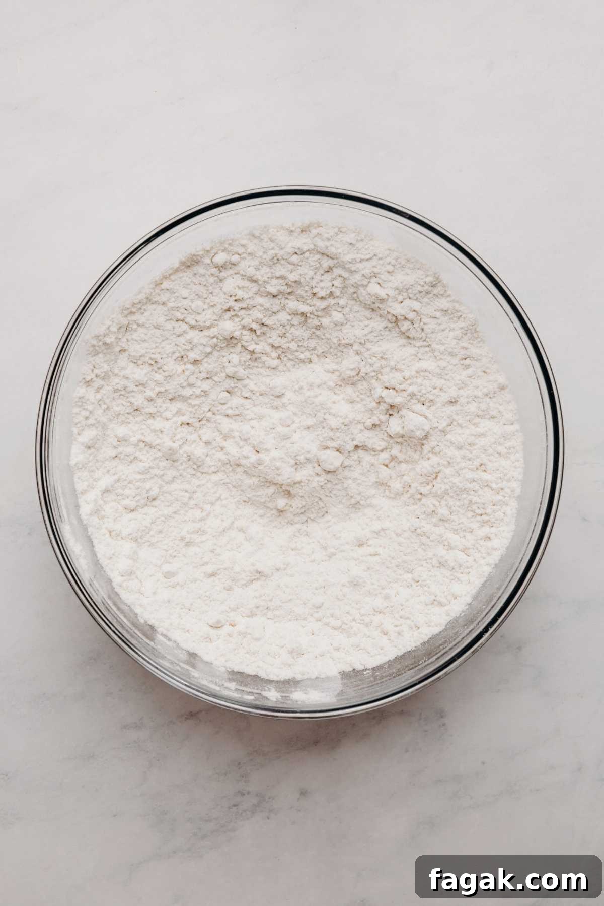 All purpose flour, baking powder, and baking soda whisked together in a large glass mixing bowl on a white marbled background.