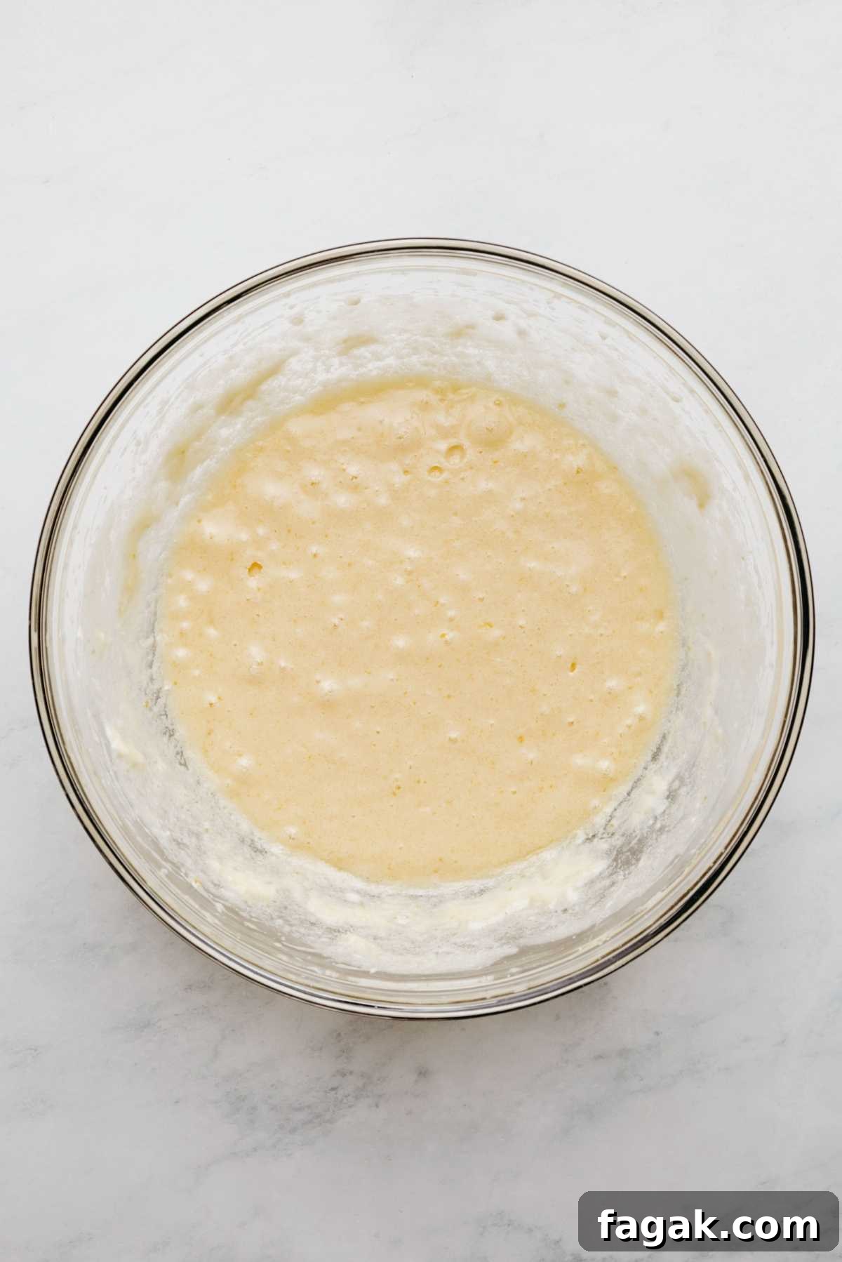 Whisked butter, sugar, eggs, and vanilla extract in a large glass mixing bowl on a white marbled background.