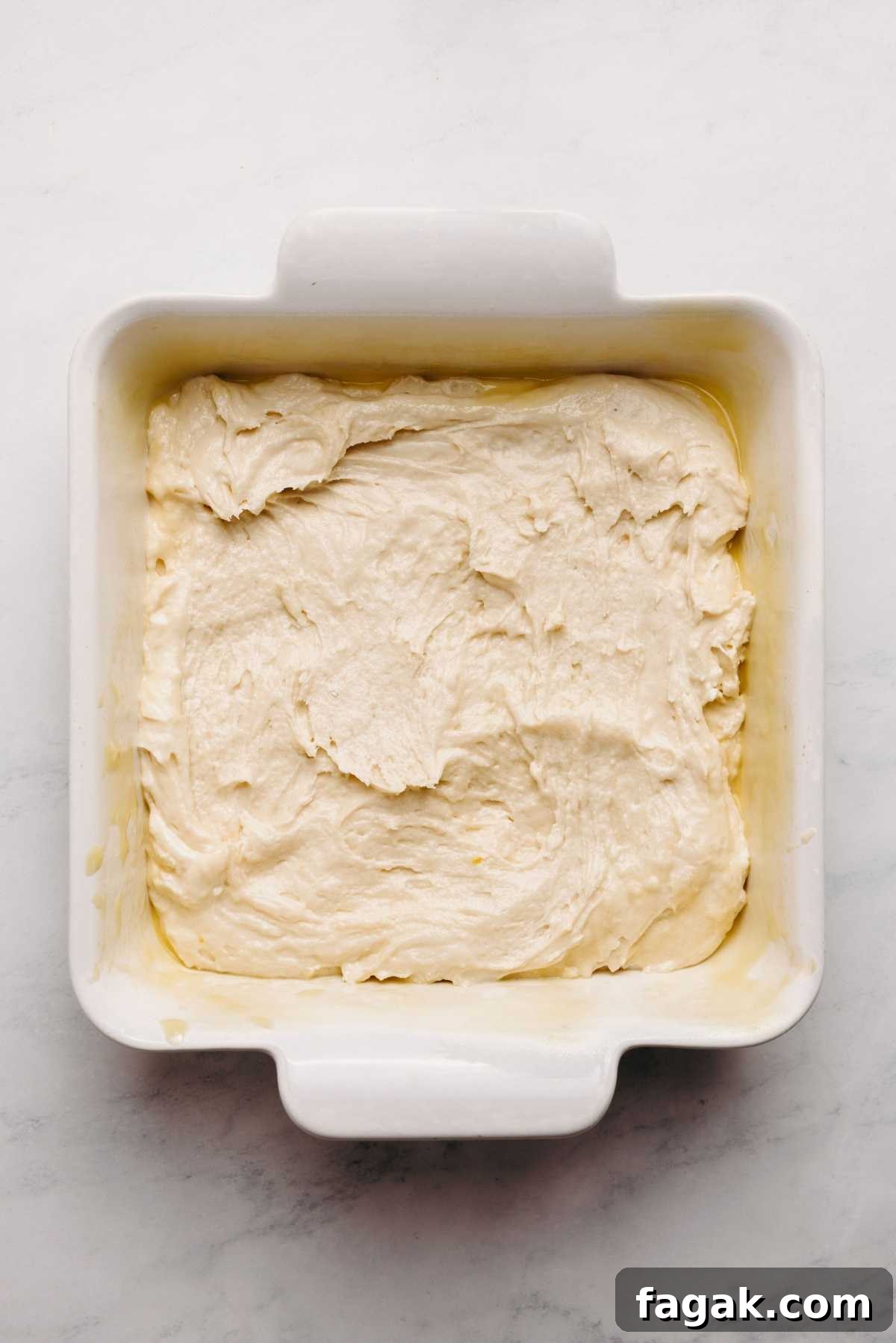 Sour cream coffee cake batter spread into a nine inch square parchment paper lined ceramic baking dish on a white marbled background.