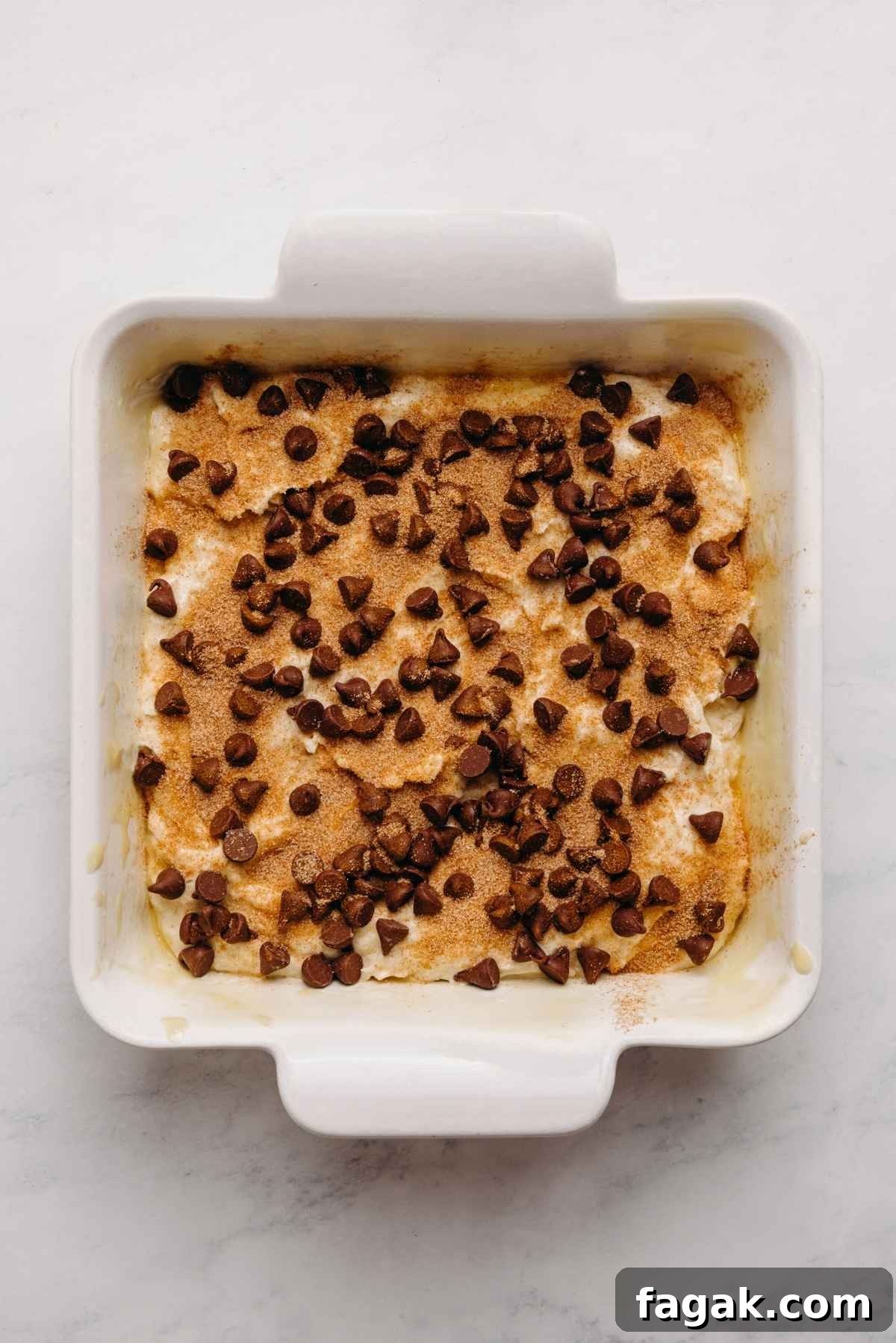 Chocolate chips and cinnamon sugar added to the top of the sour cream coffee cake batter in a nine inch square parchment paper lined ceramic baking dish on a white marbled background.