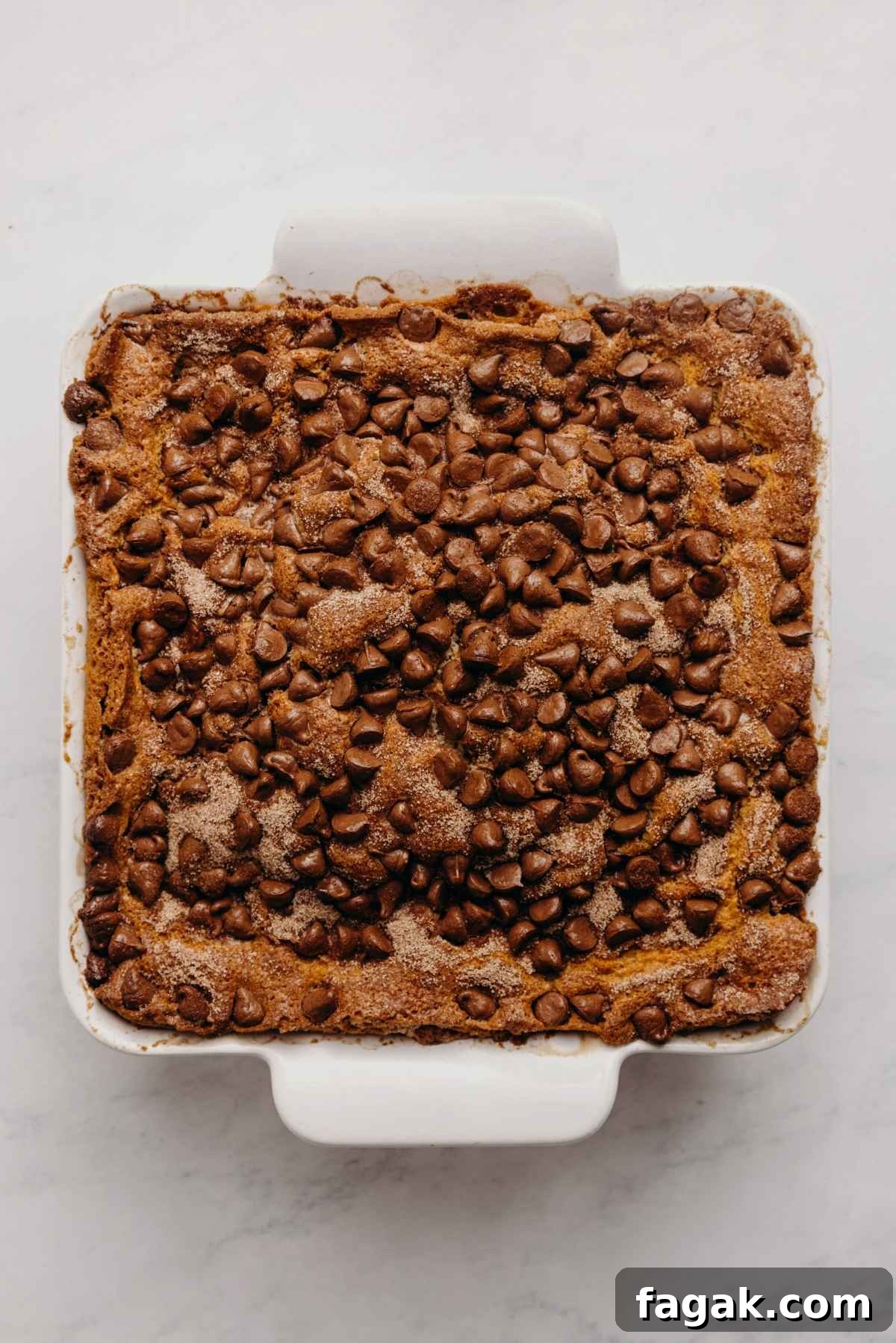 Baked chocolate chip sour cream coffee cake in a nine inch square parchment paper lined ceramic baking dish on a white marbled background.