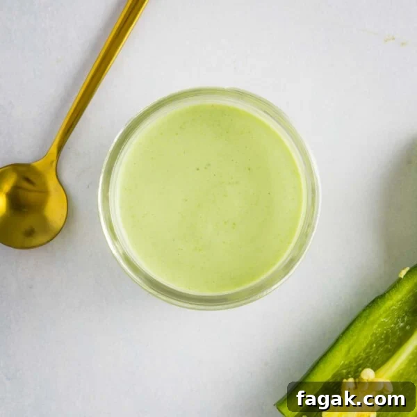 An overhead image of jalapeno ranch dressing in a glass bowl with a golden spoon on the upper left side and a jalapeno on the lower right side.