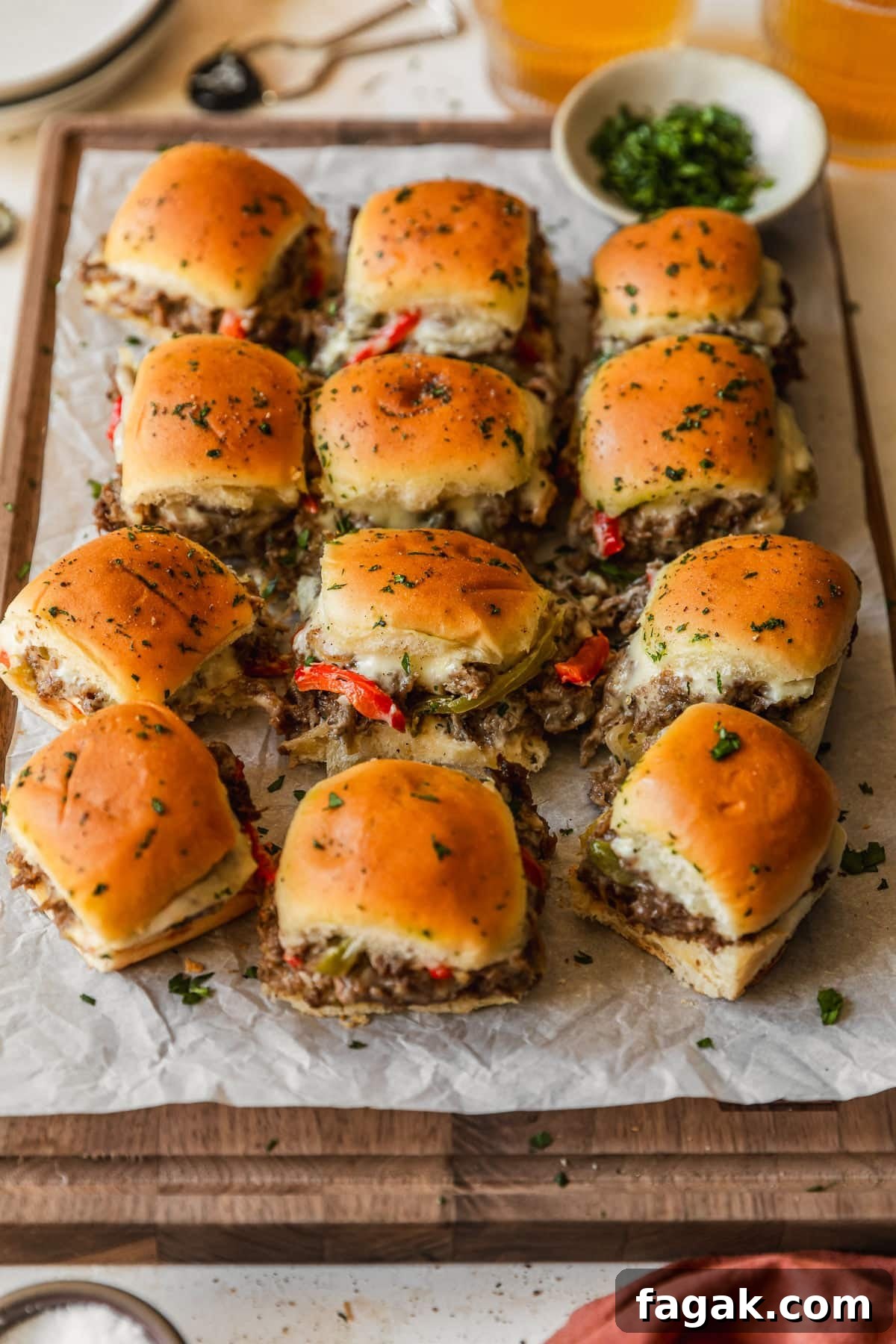 Rows of Philly cheesesteak sliders with Hawaiian rolls on a wood board next to glasses of beer, a white bowl of parsley, and an orange linen.