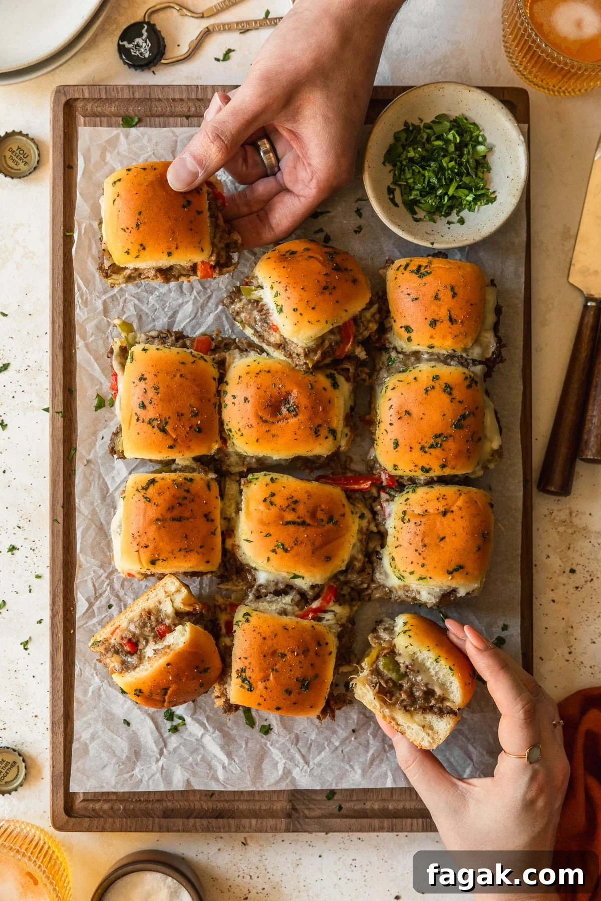 Hands reaching for Philly cheesesteak sliders on a wood board next to glasses of beer, white plates, and a beer opener with a tan background.