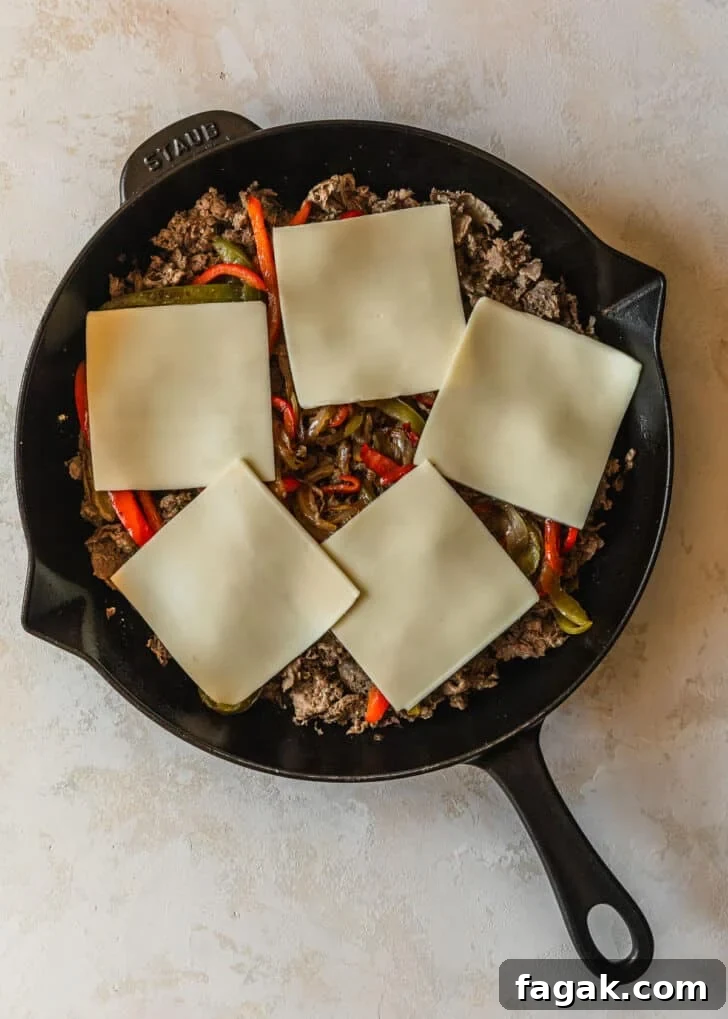 A cast iron skillet with meat, veggies, and cheese slices on a tan background.