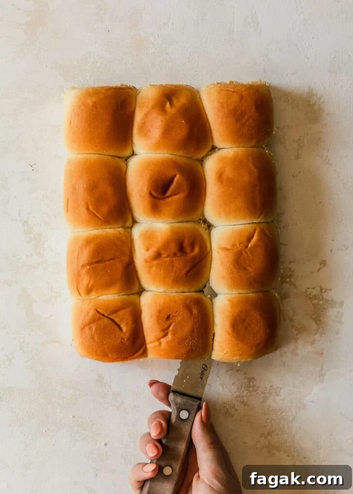 A hand using a knife to slice Hawaiian rolls in half on a tan backdrop.