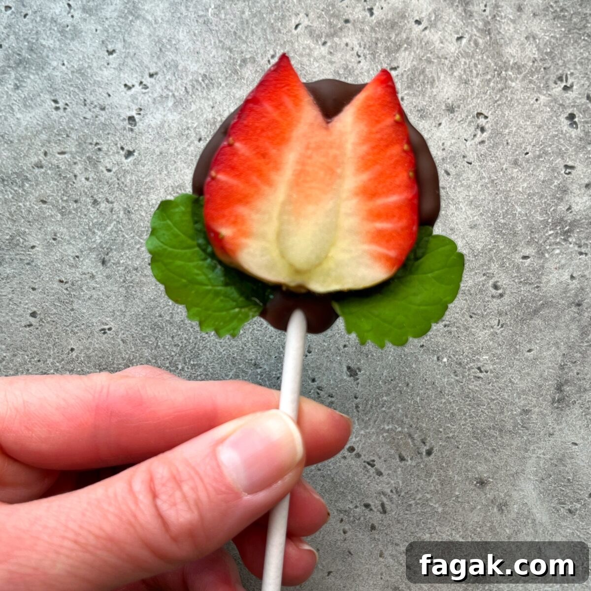 A hand holds a lollipop shaped like a tulip, made from a halved strawberry dipped in chocolate with two green leaves at the base—a creative bite perfect for health-conscious food recipes, against a gray textured background.