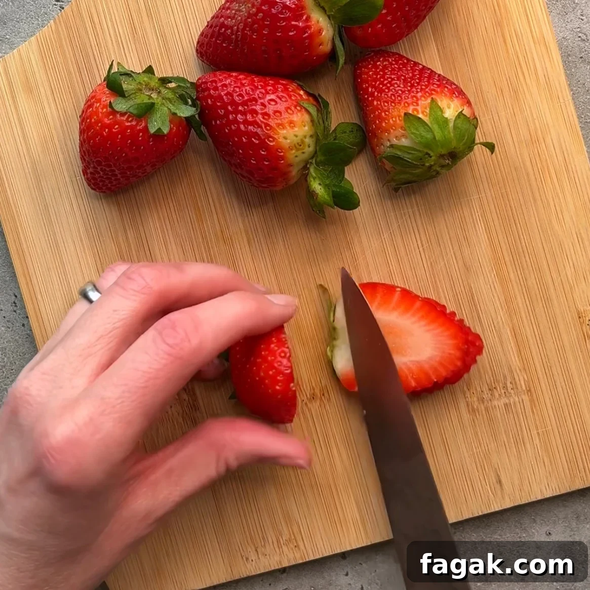 A hand slices a strawberry with a knife on a wooden cutting board.