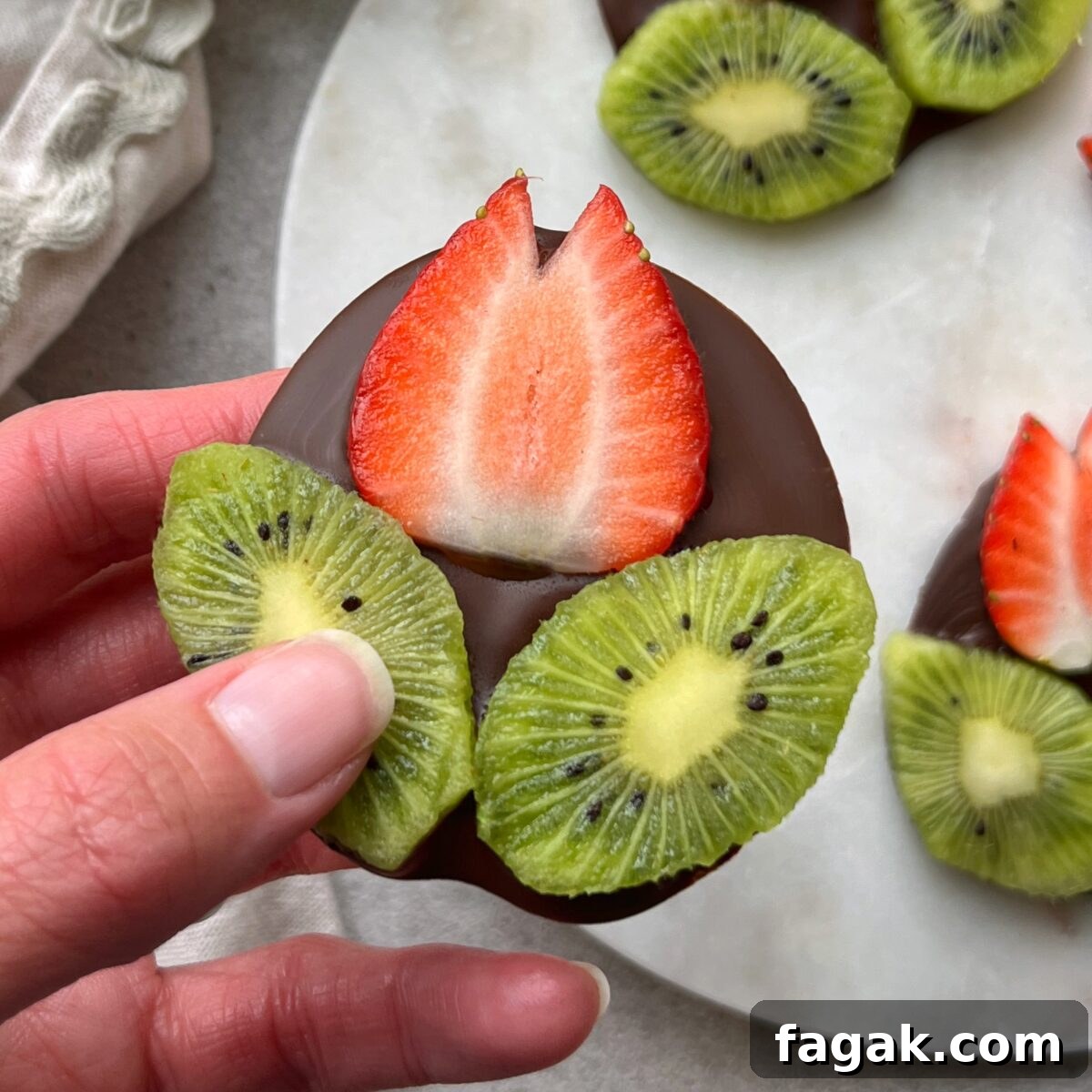 A hand holding a chocolate-covered disc topped with a slice of strawberry shaped into a flower and two slices of kiwi shaped into leaves.