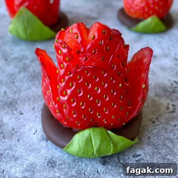 A chocolate strawberry rose, carved to resemble a blooming flower, sits atop a round piece of chocolate with a green leaf at the base, displayed on a gray surface. Other chocolate strawberry roses are softly blurred in the background.