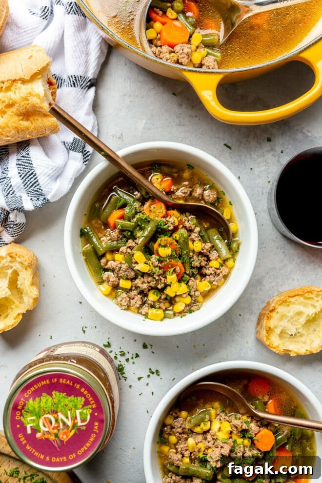 Top down of a white bowl with beef and vegetable soup on a grey surface. There is a jar of bone broth and a yellow dutch oven with soup also on the surface. There are slices of French bread with butter. 