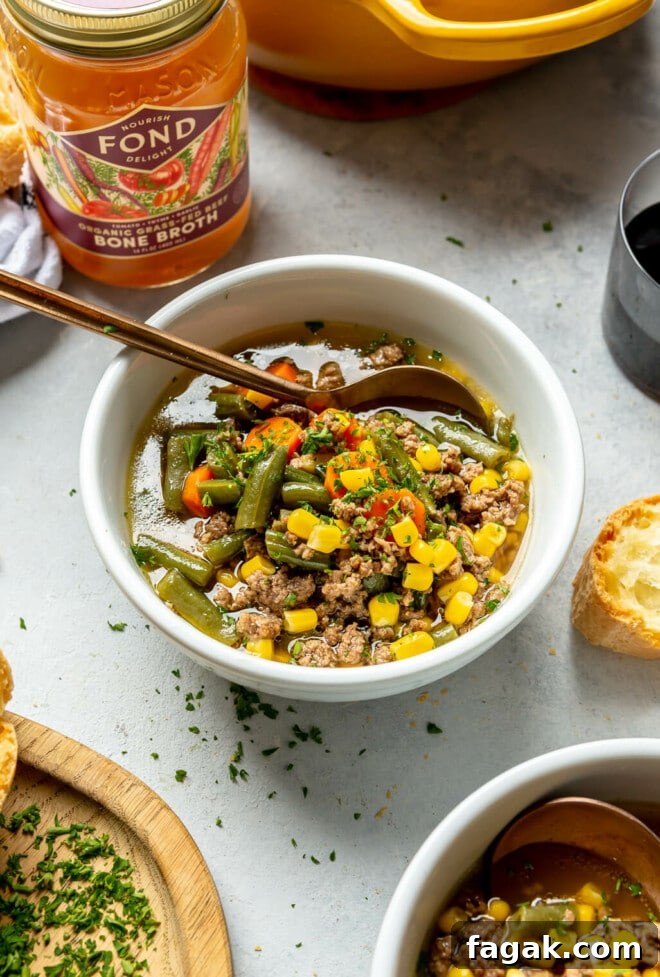 Side view of beef and vegetable soup in a white bowl with a copper spoon. There is a jar of FOND bone broth in the background. 