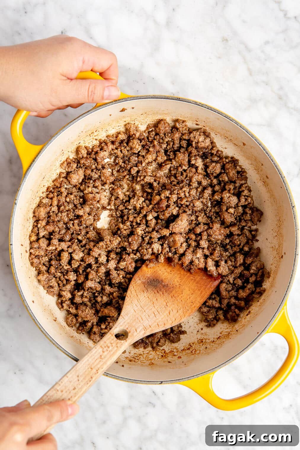 Cooked ground beef in a yellow enameled dutch oven. A hand is stirring the beef with a wooden spatula.