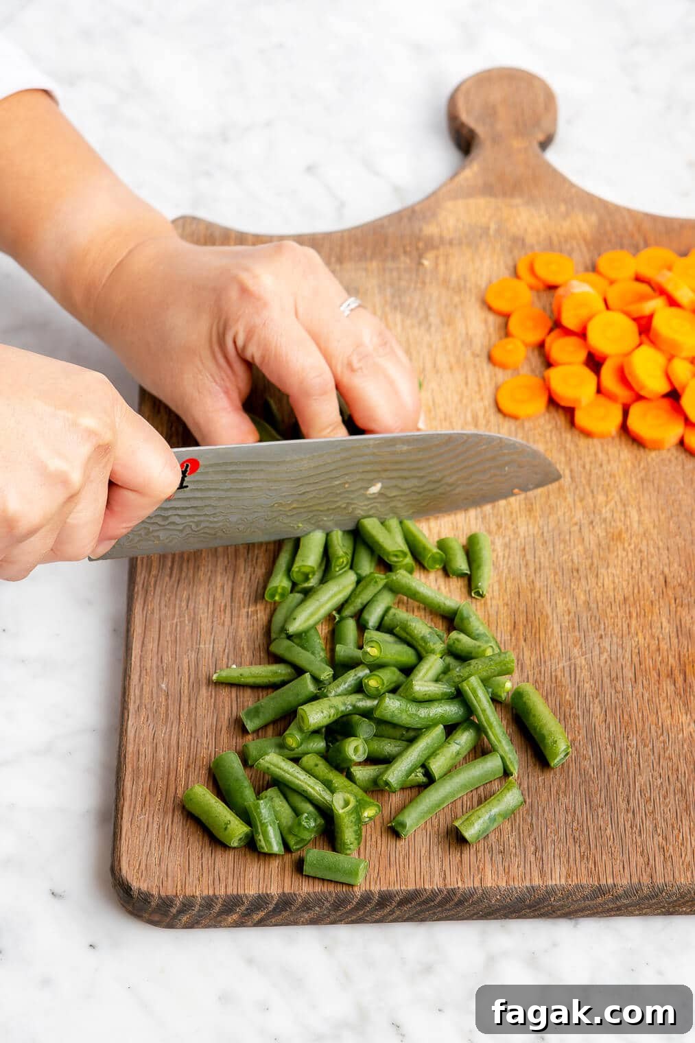 Hand holding a butcher knife to cut green beans. There are sliced carrots in the background on a wooden cutting board.