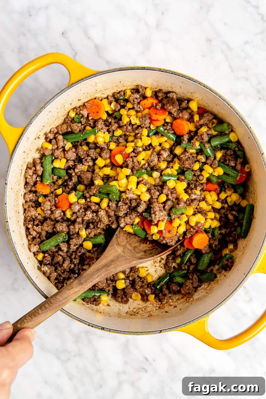Ground beef, carrots, corn, and green beans in an enameled dutch oven with a wooden spoon stirring them.