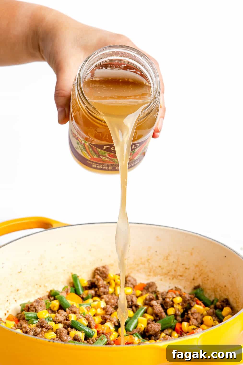 Hand pouring bone broth into the beef and vegetable mixture.