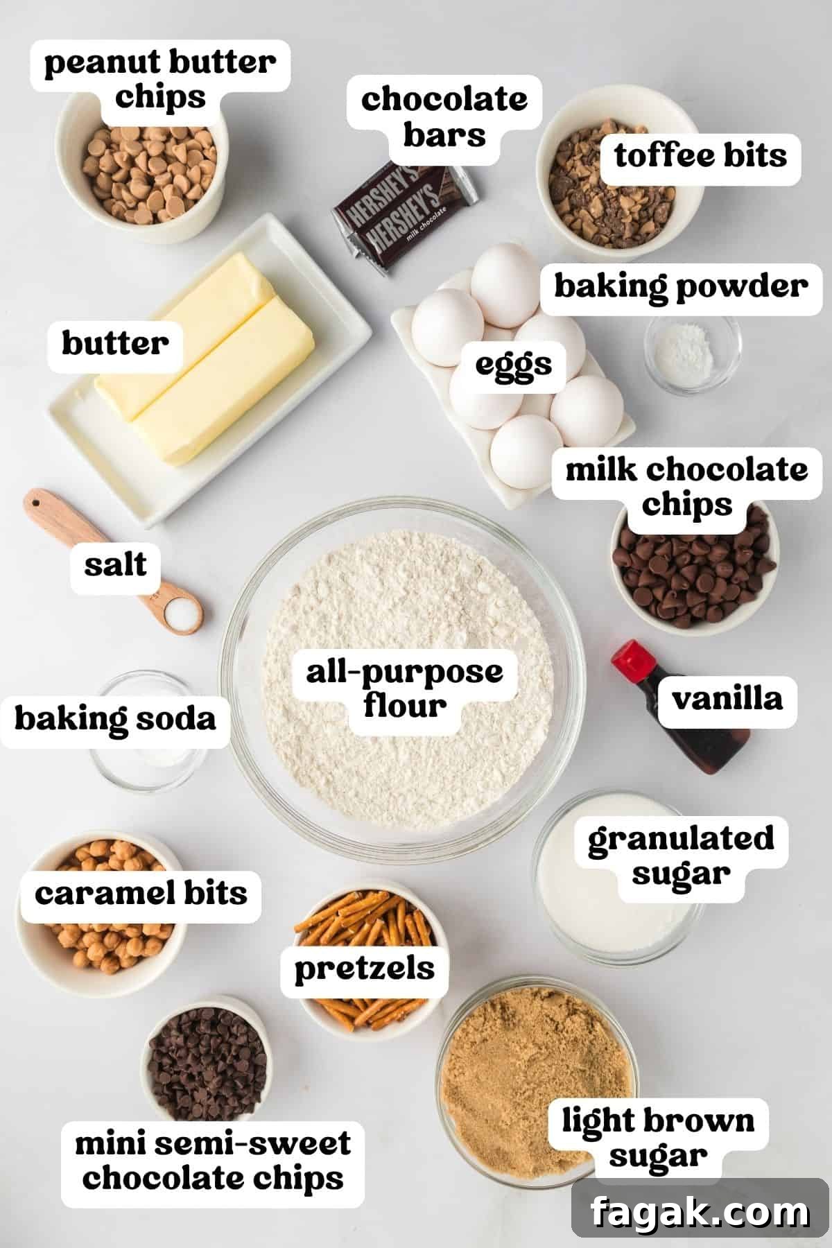 Kitchen sink cookie ingredients on countertop: peanut butter chips, chocolate bars, toffee bits, two sticks of butter, eggs, baking powder, salt, flour, milk chocolate chips, vanilla, baking soda, pretzels, granulates sugar, light brown sugar, caramel bits, and mini semi-sweet chocolate chips.