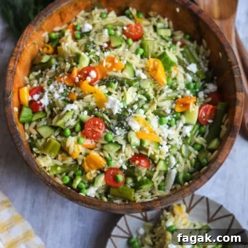 Brown bowl of orzo asparagus salad with orzo pasta, grilled asparagus and bell peppers, peas, cucumbers, vegan feta cheese and cherry tomatoes with a Dijon mustard vinaigrette dressing, with a serving of the salad on a separate plate.