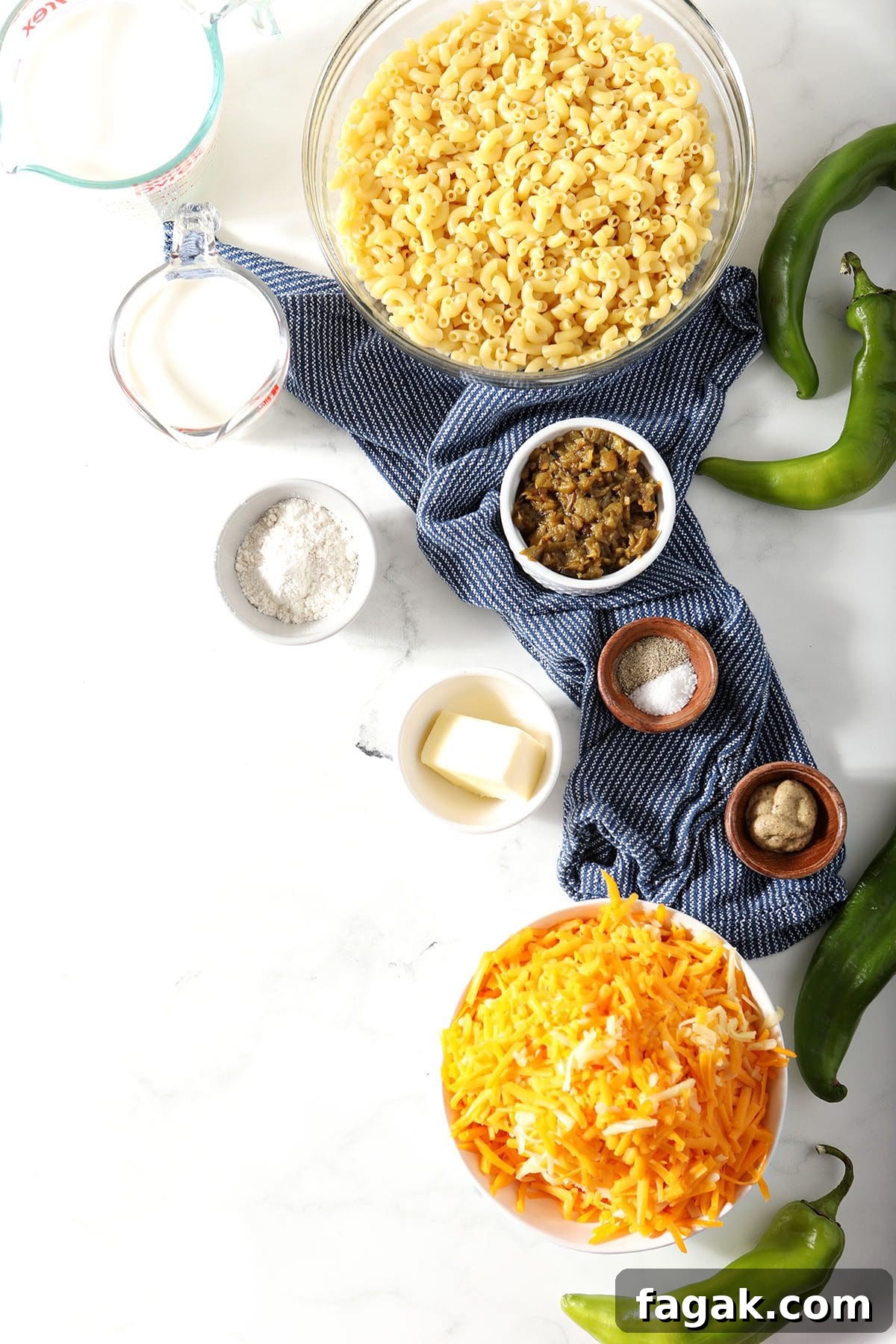 Ingredients to make macaroni and cheese in bowls on marble surrounded by green Chile peppers