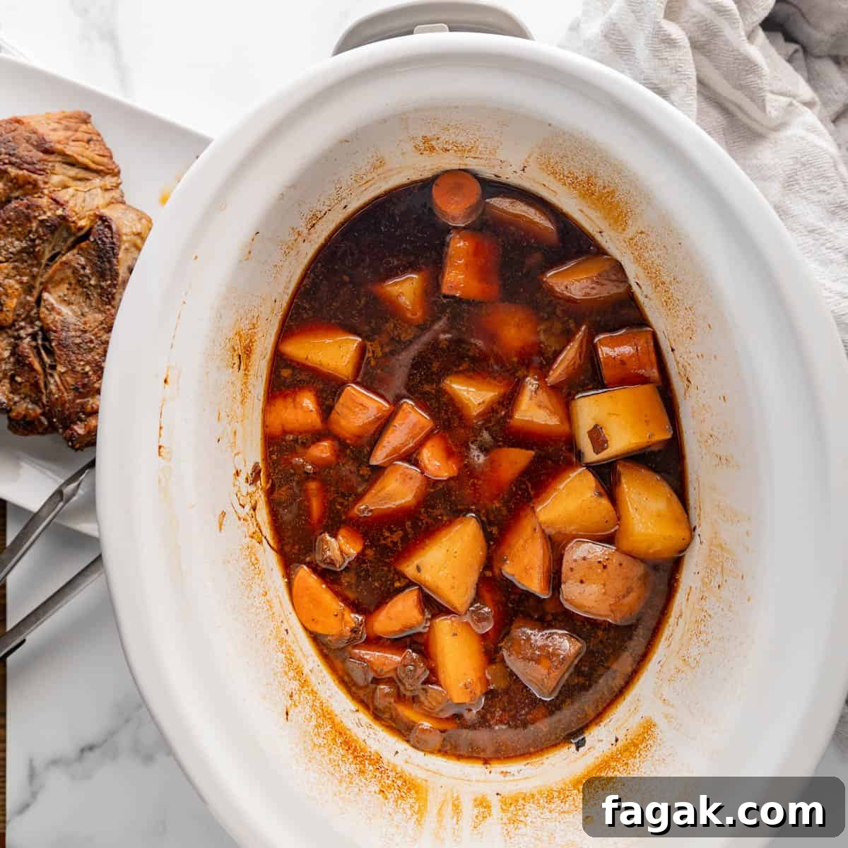 Slow cooker with a stew of beef, potatoes, and carrots in a rich broth, next to a plate with cooked meat on a white table.