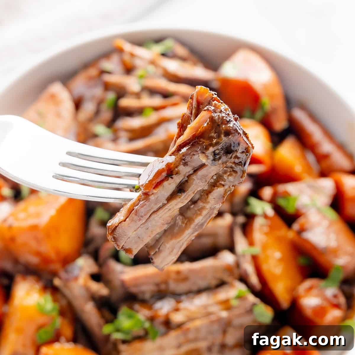 A fork holding a piece of cooked meat with a bowl of meat and roasted vegetables in the background.