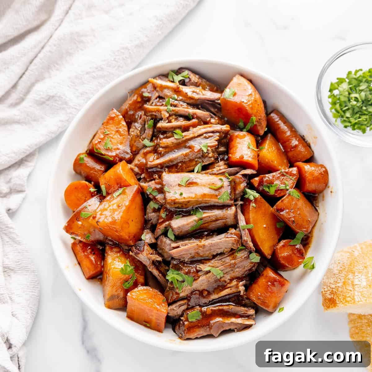A bowl of pot roast with slices of beef, potatoes, and carrots in gravy, garnished with herbs. There's a side of bread and a small dish of chopped herbs.