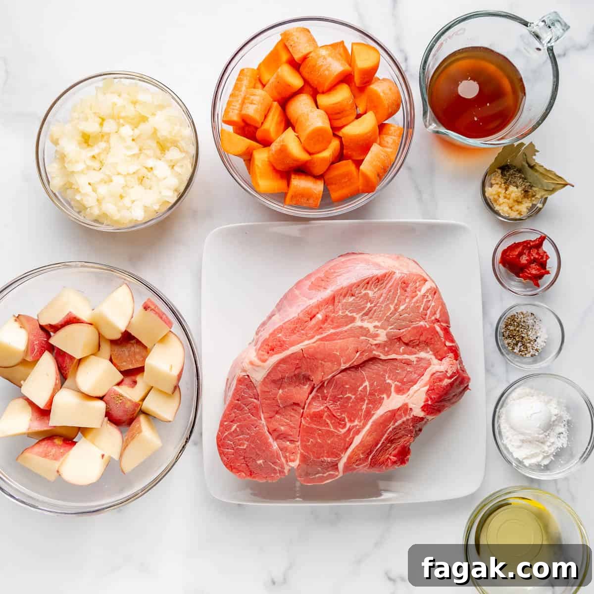 Raw beef roast on a plate, surrounded by bowls of chopped carrots, potatoes, onions, beef broth, minced garlic, bay leaves, tomato paste, spices, cornstarch, and oil.