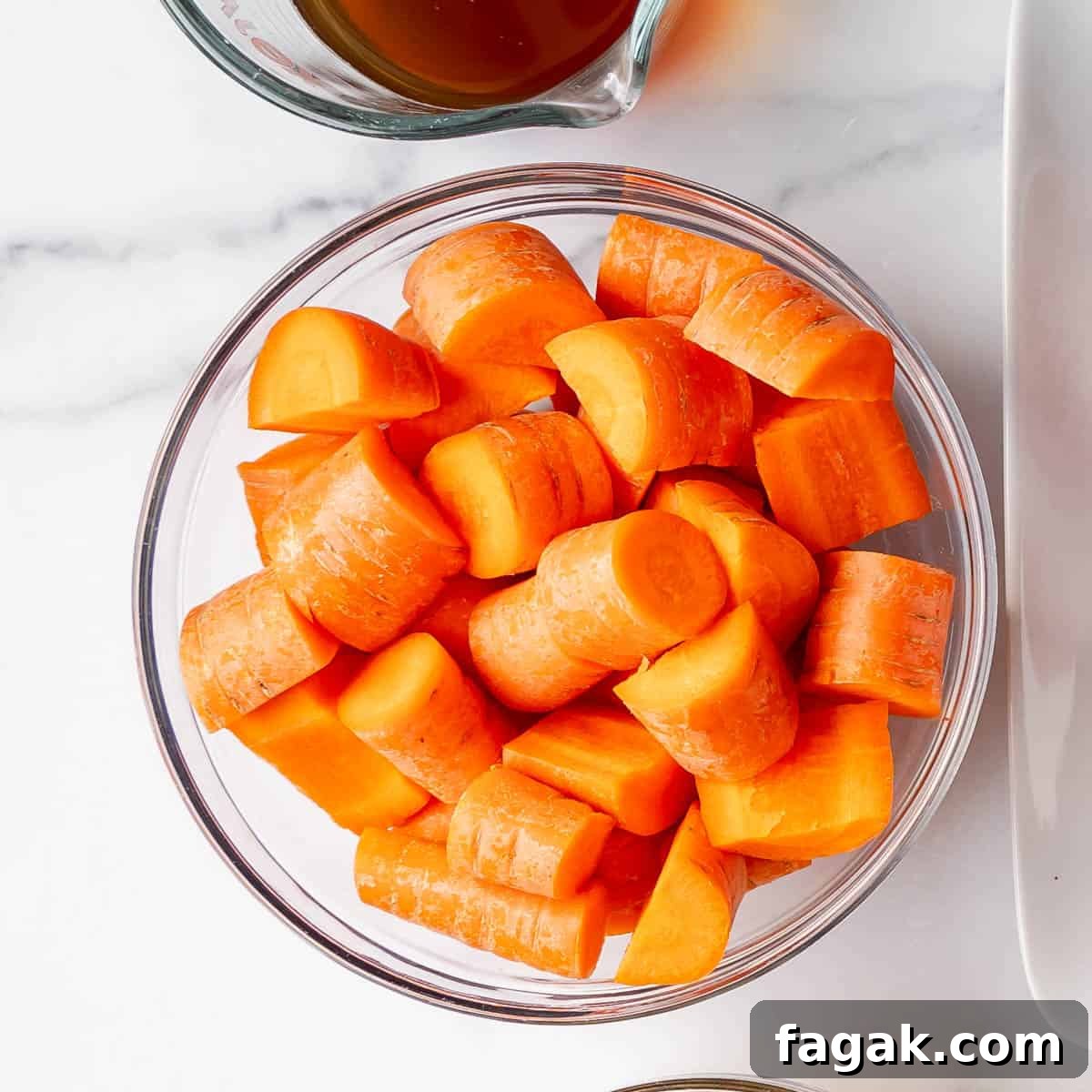 A glass bowl filled with peeled, sliced carrots on a white surface, ready to complement a hearty crockpot pot roast. Nearby, part of a clear container with savory liquid is visible.