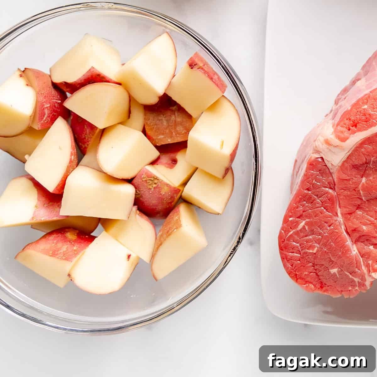A bowl of cubed red potatoes sits beside a raw piece of beef, ready to transform into a hearty Crockpot Pot Roast on the pristine white surface.