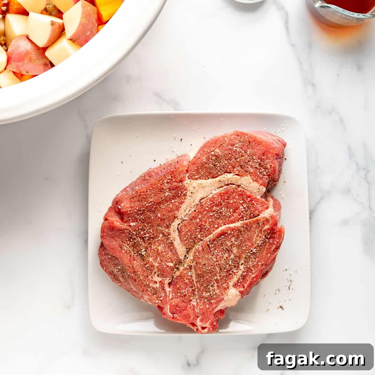A raw, seasoned beef steak rests on a square white plate. Nearby are chopped vegetables in a bowl on a marble surface.