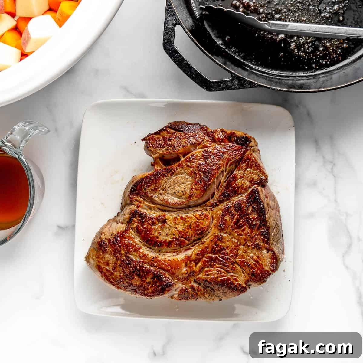 A cooked piece of steak on a white square plate, next to a metal pan, a glass pitcher, and a bowl with chopped vegetables on a marble countertop.