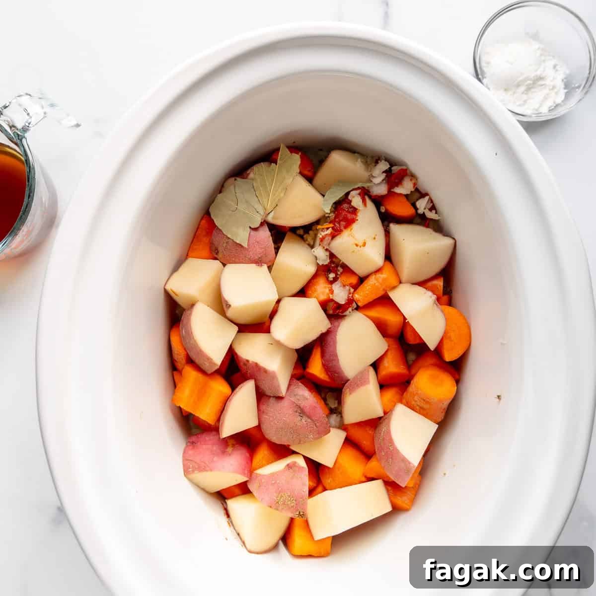 A slow cooker filled with chopped potatoes, carrots, and spices. A glass measuring cup with broth and a small bowl of white powder are nearby on a marble surface.