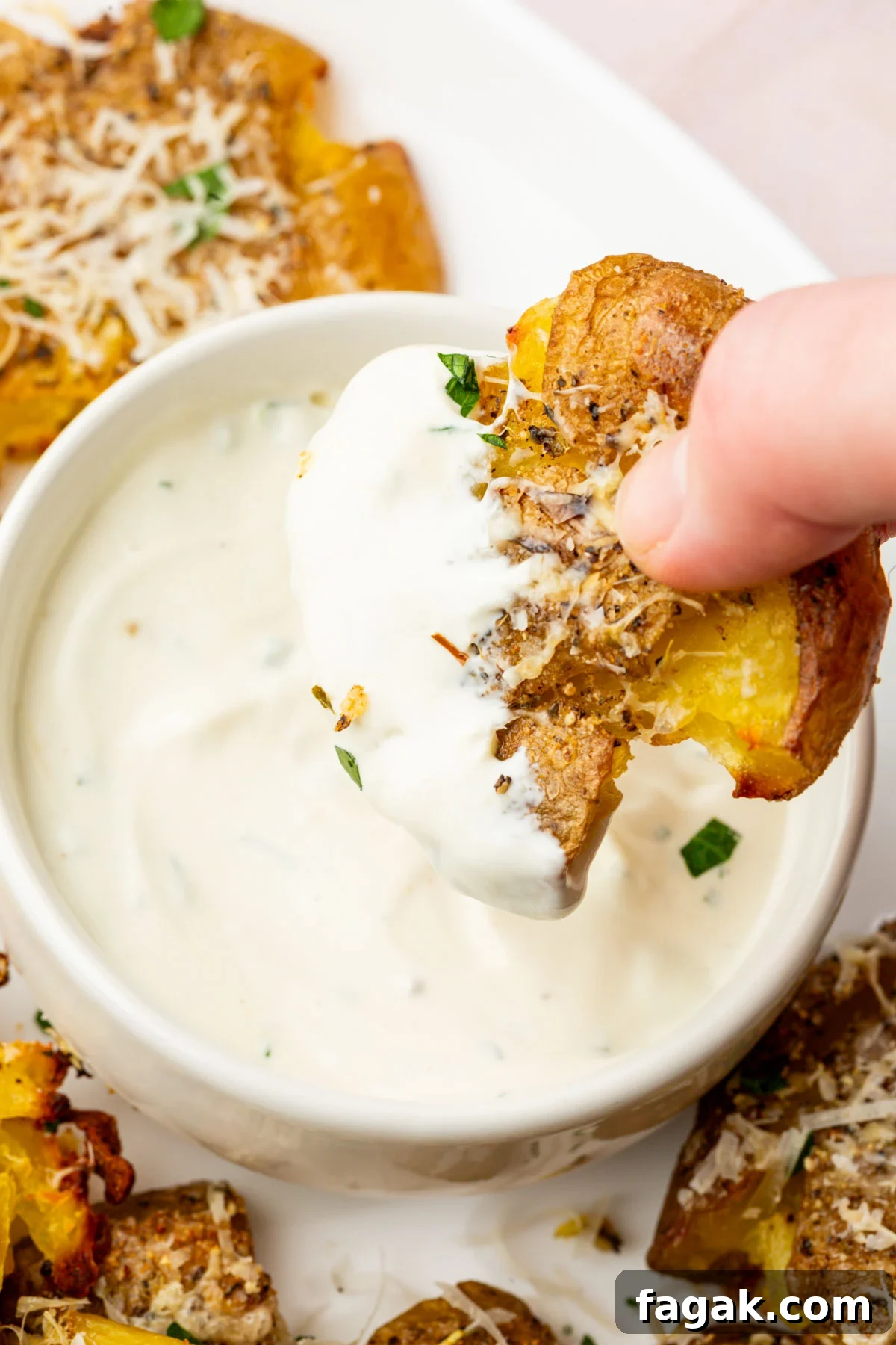 Hand dipping a cheesy smashed potato into a bowl of creamy white sauce.