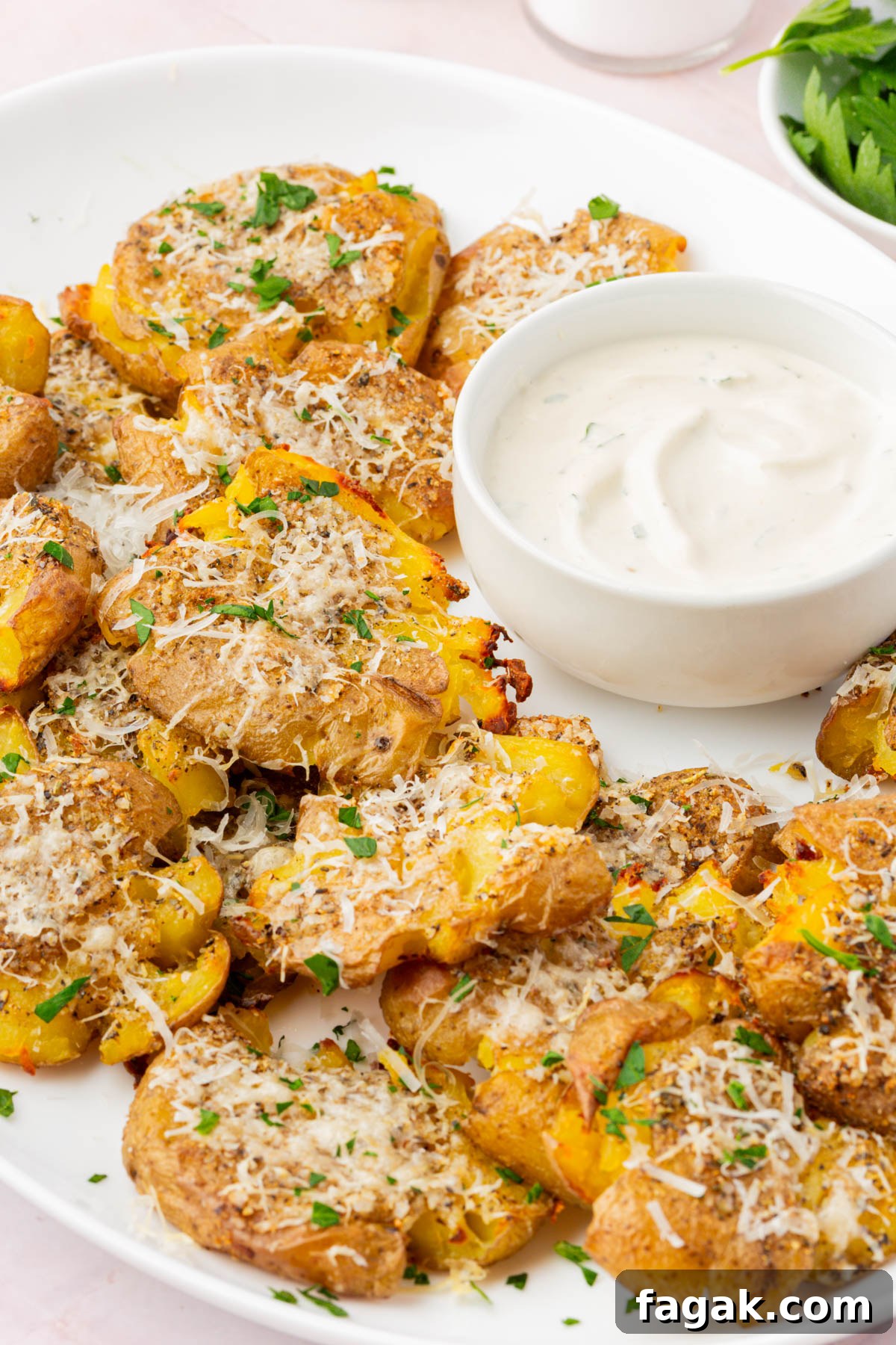 Crispy parmesan smashed potatoes served on a white platter with a bowl of creamy dipping sauce.
