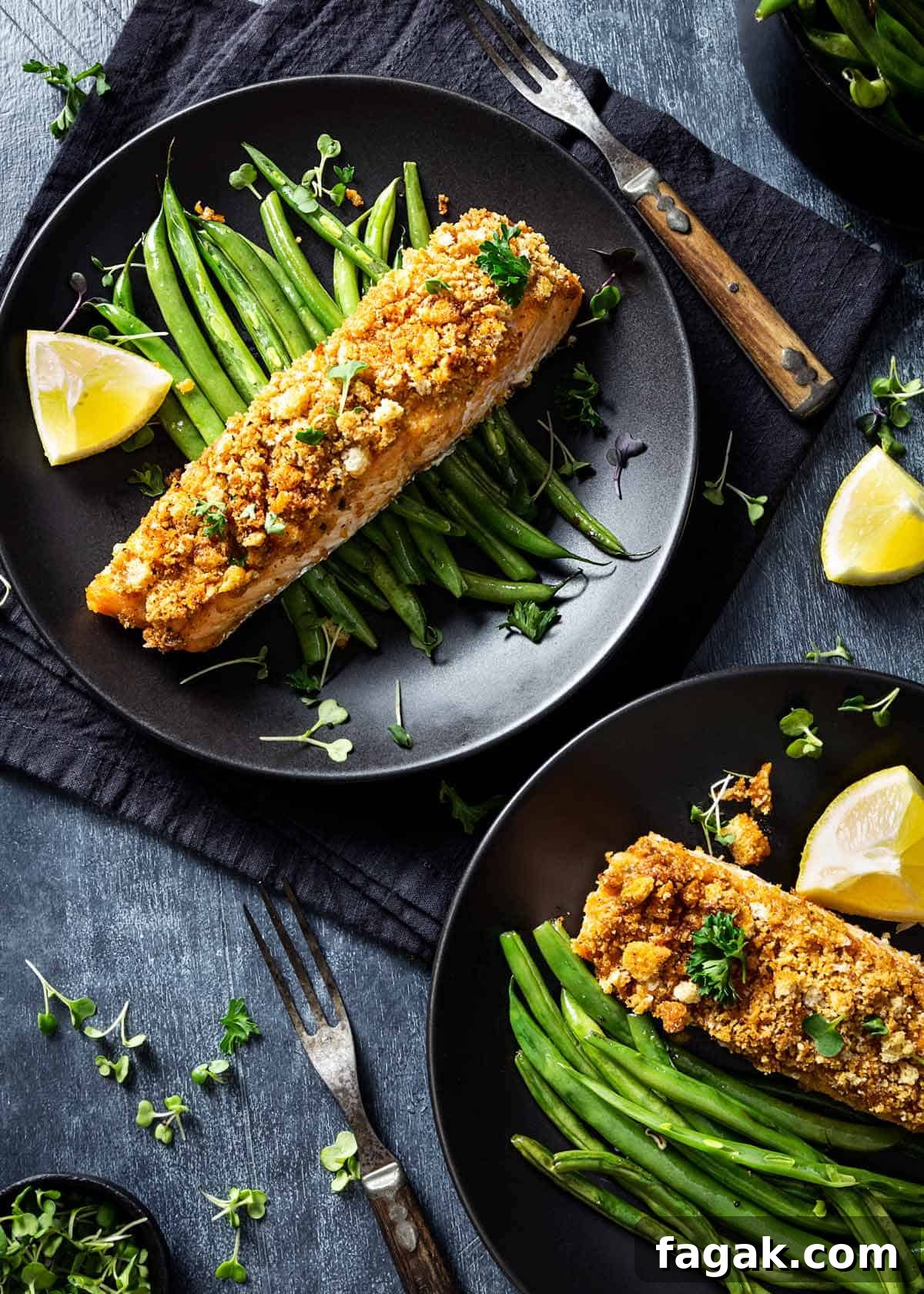 Two plated salmon filets over green beans with lemon wedges on a table.