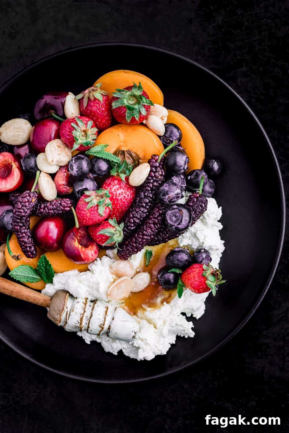Summer fruit with fresh cheese, almonds and honey on a black plate; overhead shot on a black background.