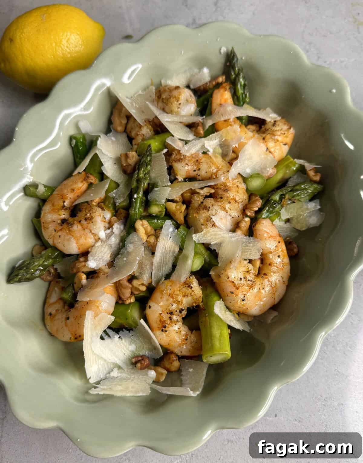 A scalloped green dish containing sautéed shrimp, asparagus, walnuts, and shaved parmesan cheese. A whole lemon sits next to the dish on a light-colored surface.
