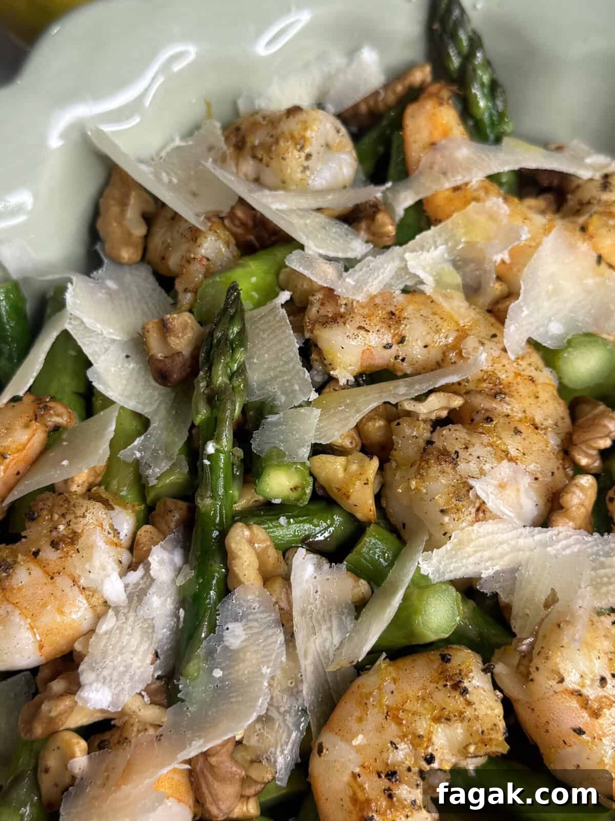 Close-up of a dish featuring cooked shrimp, fresh asparagus, walnut pieces, and shaved Parmesan cheese, all seasoned with black pepper and served on a light green plate.