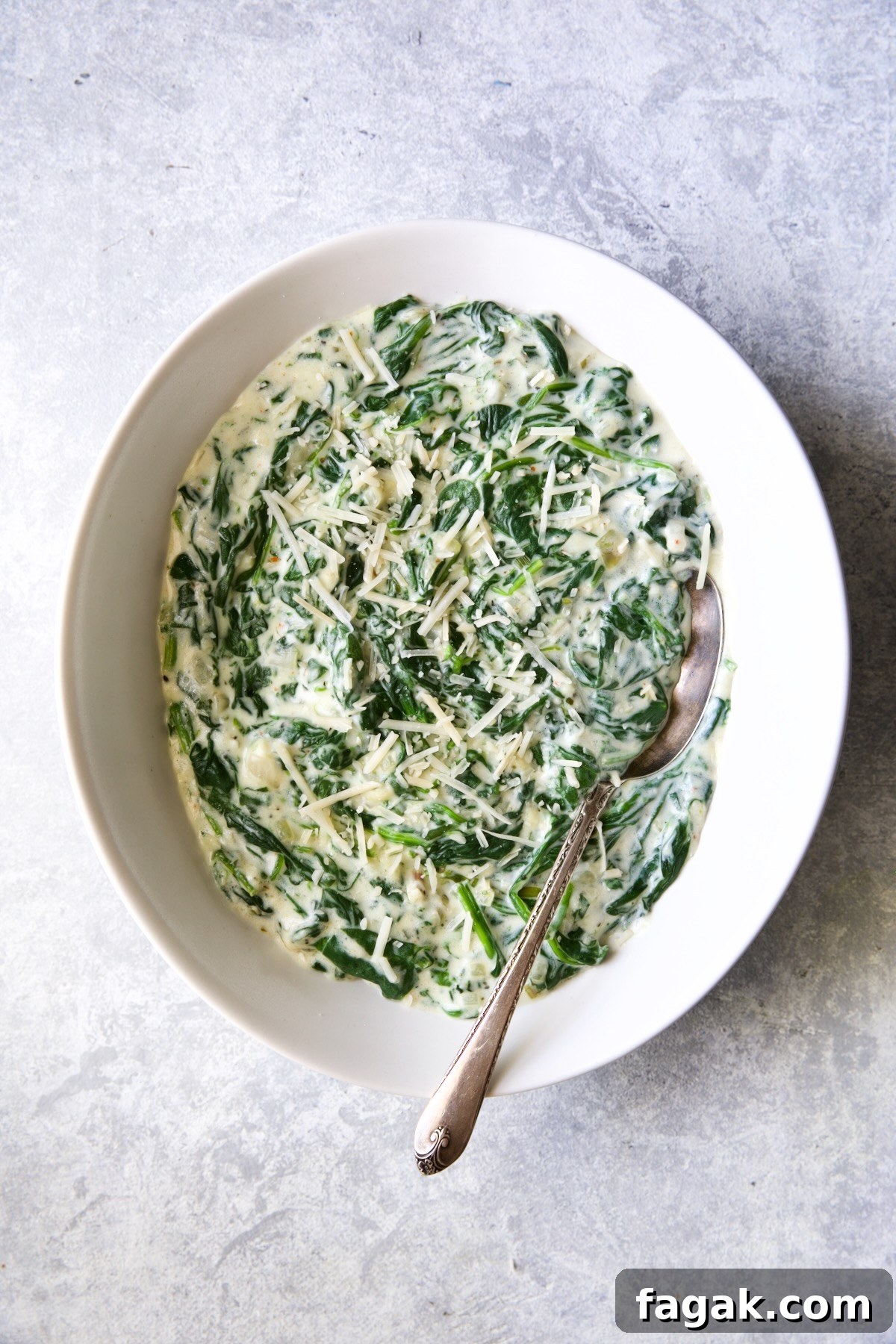 Steakhouse Creamed Spinach in white oval bowl with serving spoon on light blue-gray background.