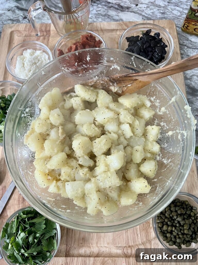 Overhead photo of potatoes mixed with lemon garlic olive oil dressing in glass bowl with wooden spoon, surrounded by the remaining prepped salad ingredients.