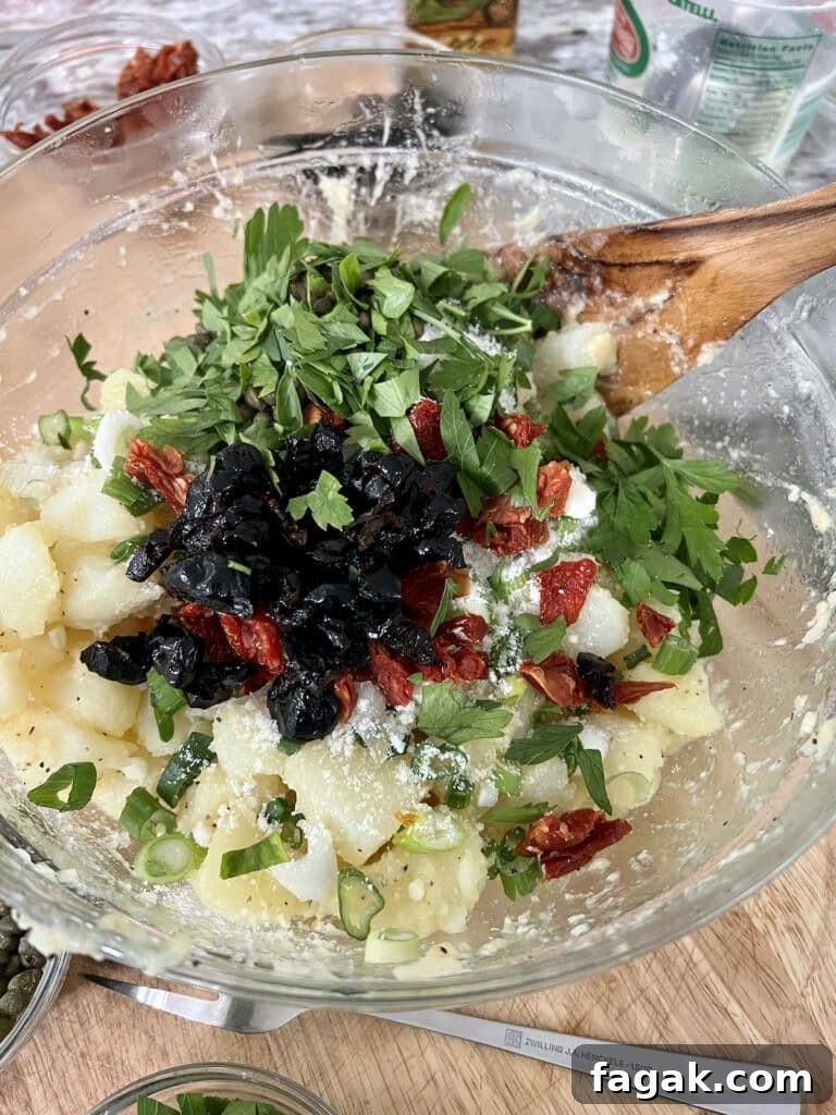 Mixing chopped parsley, black olives and sun dried tomatoes into potato salad with wooden spoon in glass bowl.