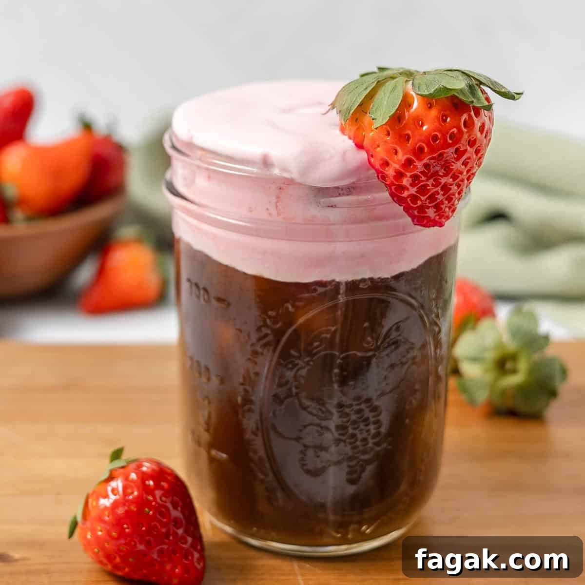 A glass jar filled with iced coffee topped with pink foam and garnished with a fresh strawberry, with more strawberries in the background.
