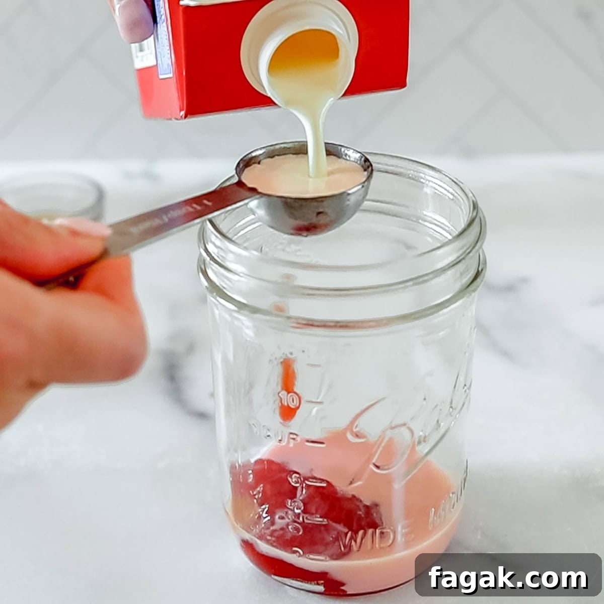A hand pours cream from a carton into a tablespoon over an empty glass jar, with some cream already inside the jar.